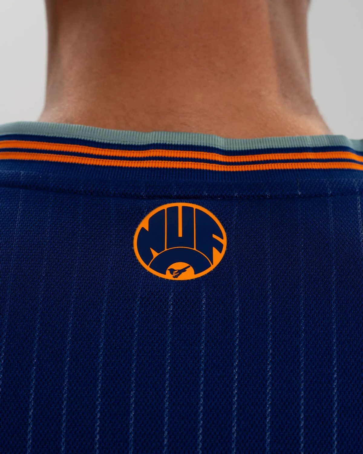 Back view of a person wearing a dark blue athletic jersey with orange stripes and a circular orange logo on the back collar area, neck visible.