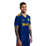 Man in a blue soccer jersey with orange accents, smiling and standing sideways, tattoos visible on both arms.