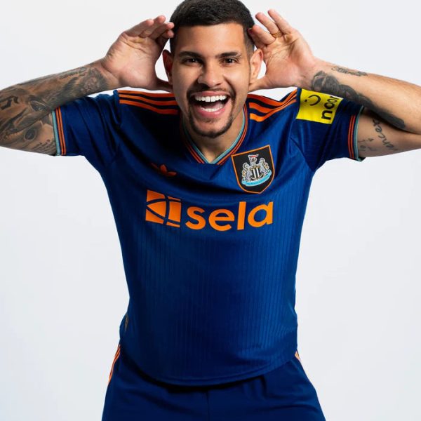 Soccer player in blue kit smiles with hands cupped behind ears, posing for the camera