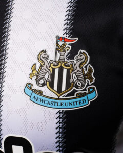 Newcastle United crest on a black-and-white striped jersey, with two seahorses supporting a shield and a blue banner reading Newcastle United.