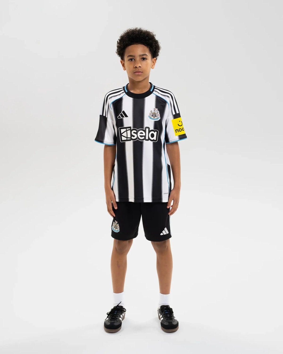 Young boy standing in a Newcastle United youth football jersey with black-and-white stripes, sponsor logo 'Isela' on chest and club crest on left side.