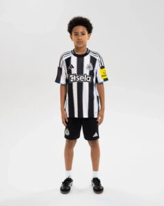 Young boy standing in a Newcastle United youth football jersey with black-and-white stripes, sponsor logo 'Isela' on chest and club crest on left side.
