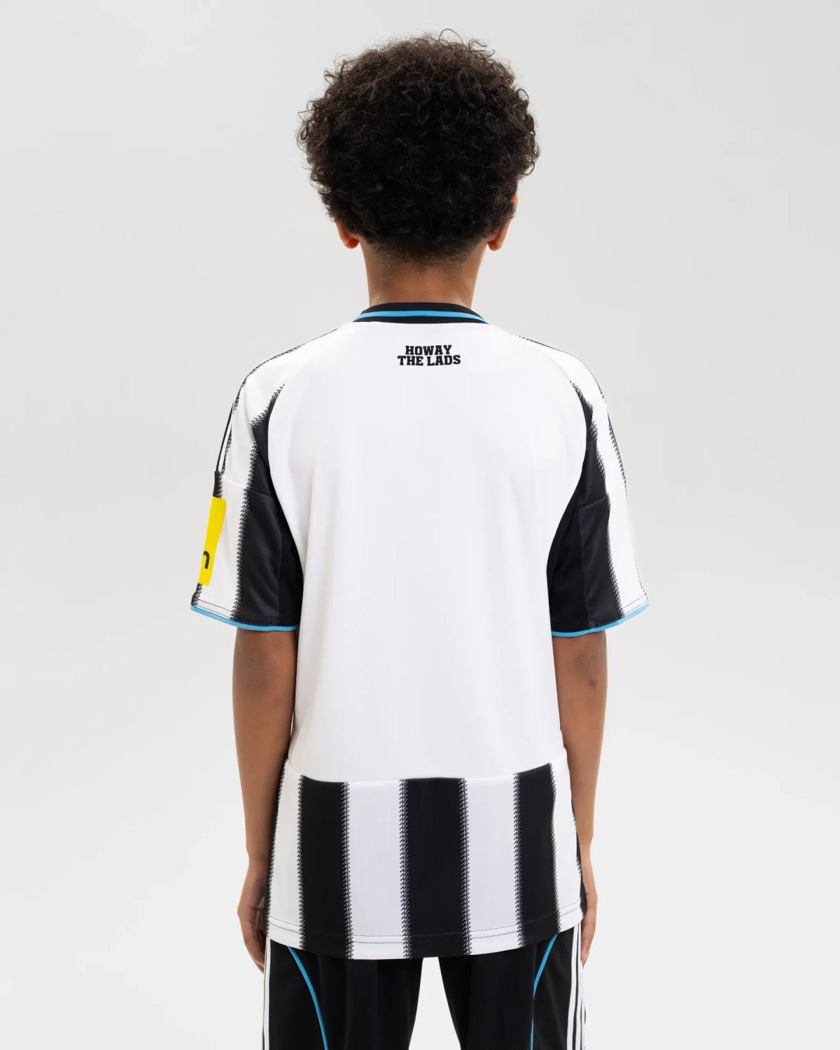 Back view of a person wearing a white soccer jersey with black vertical stripes and blue trim; 'HOWAY THE LADS' is printed on the upper back.