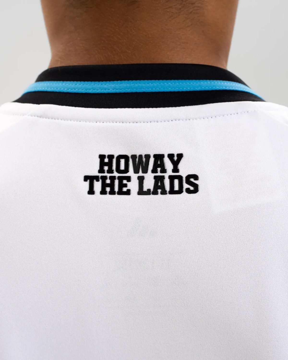 Back of a person wearing a white shirt with a turquoise collar and the text 'HOWAY THE LADS' visible on the back edge.