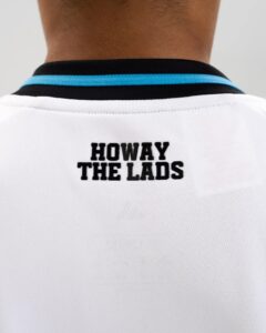 Back of a person wearing a white shirt with a turquoise collar and the text 'HOWAY THE LADS' visible on the back edge.