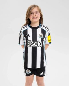Smiling child wearing a Newcastle United black-and-white striped soccer jersey with a yellow armband, standing against a light gray background.