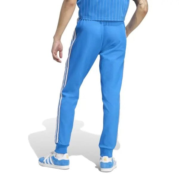 Back view of a person wearing bright blue athletic pants with white side stripes and matching blue sneakers, standing casually on a white background.