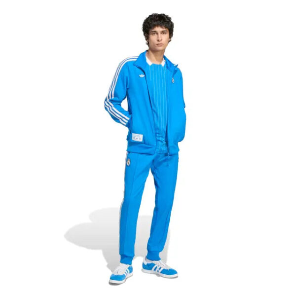 Young man standing in a bright blue tracksuit with white stripes, hands in pockets, white sneakers, on a white background.