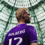 Side profile of a soccer player in a purple jersey with the name Dolberg and number 12 on the back, standing in a glass-domed stadium.
