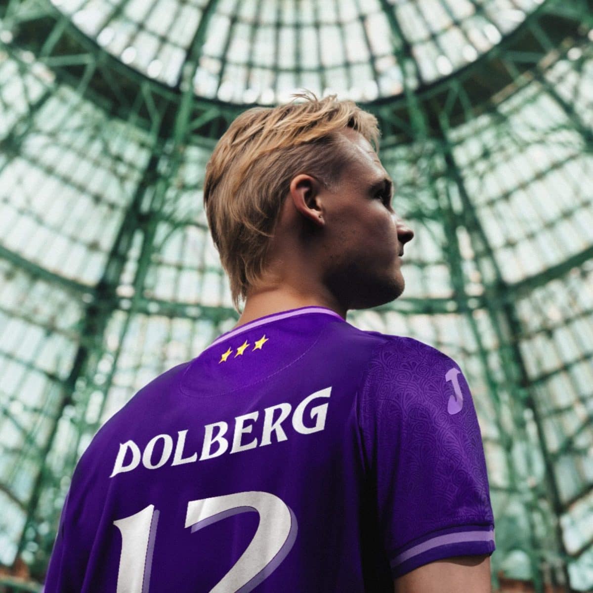 Back view of a soccer player in a purple Dolberg jersey with the number 12, standing inside a glass-domed arena featuring green metal framework.