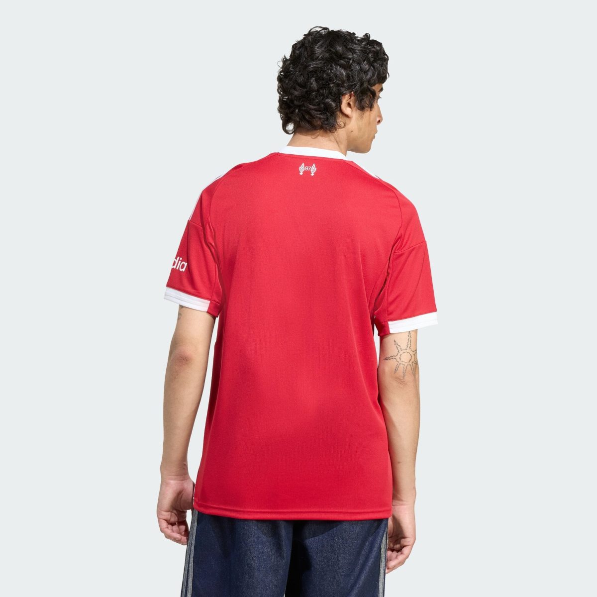 Person wearing a red sports jersey photographed from behind, showing the back of the shirt with white trim and a visible forearm tattoo, against a light gray background.