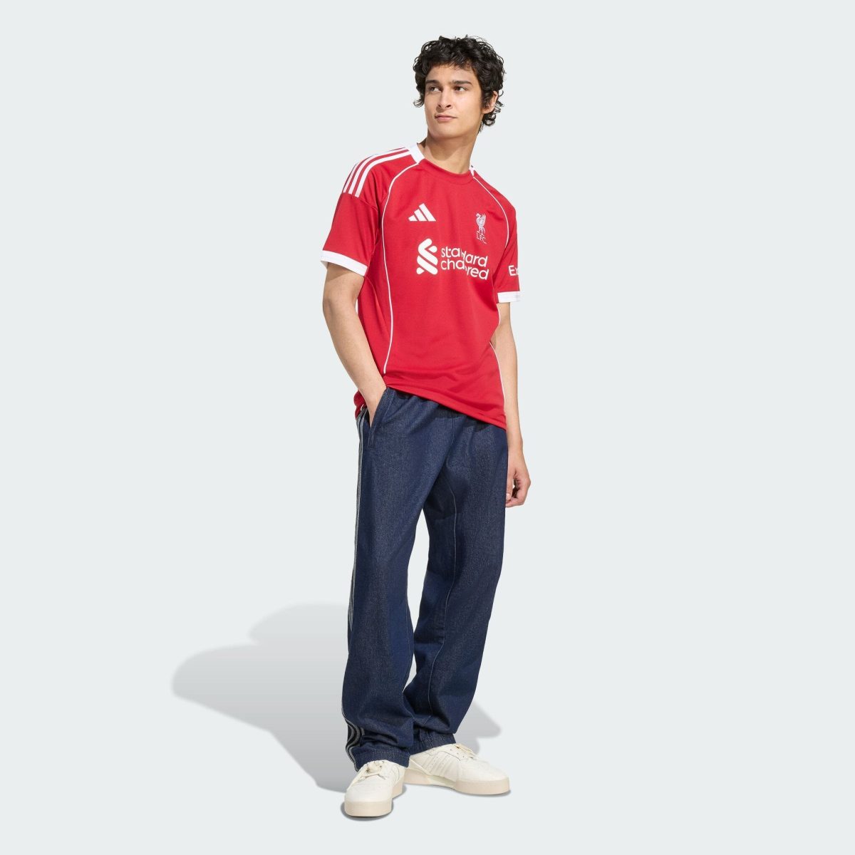 Young man in a red Liverpool FC jersey and dark jeans, standing with one hand in his pocket against a light gray background