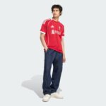 Young man in a red Liverpool FC jersey and dark jeans, standing with one hand in his pocket against a light gray background