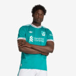 Man wearing a teal Liverpool FC jersey with the Standard Chartered sponsor, arms crossed, looking at the camera.