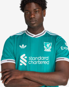 JV6427_7_APPAREL_OnModel_DetailView1_custom_1-lowres | UAE SPORT STORE Young man with arms crossed wearing a teal Liverpool FC jersey featuring Standard Chartered and Adidas logos.