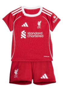 Red Liverpool FC football jersey with white Adidas stripes on the shoulders, crest, and sponsor logos (Standard Chartered) visible; paired with matching red shorts showing club emblem.