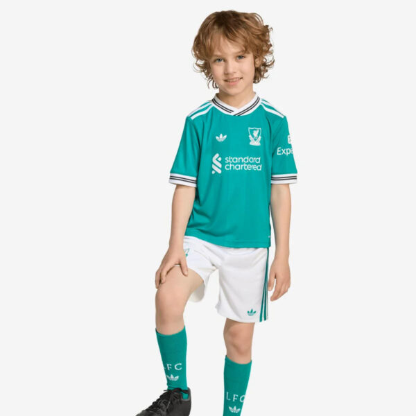 Young boy posing in a teal Liverpool FC jersey and white shorts, one knee raised, smiling at the camera.