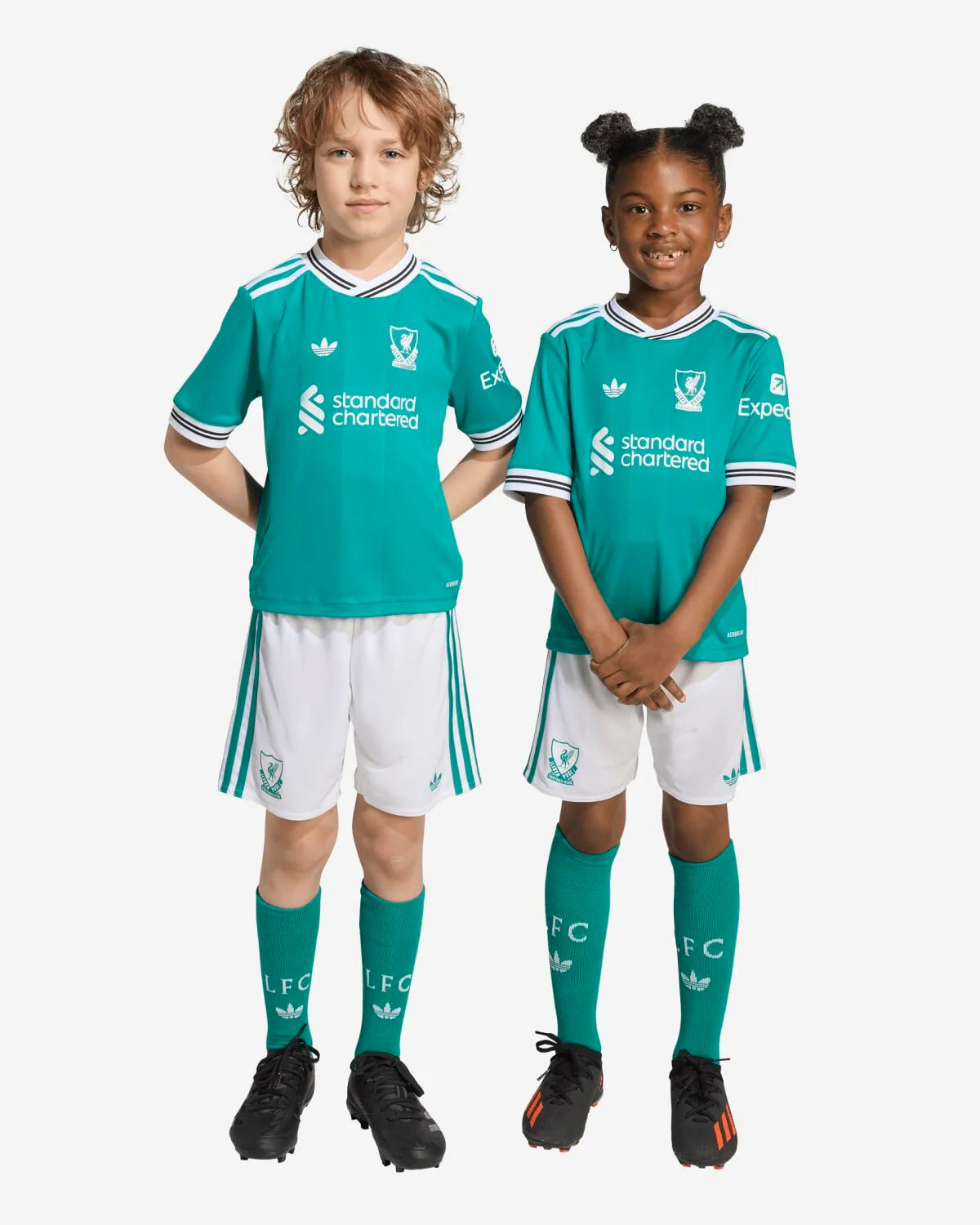 JV6470_8_APPAREL_OnModel_StandardUnisexPairShot_custom_1-lowres | UAE SPORT STORE Two children wearing teal Liverpool FC youth soccer jerseys and white shorts, teal socks, and black cleats, standing side by side.