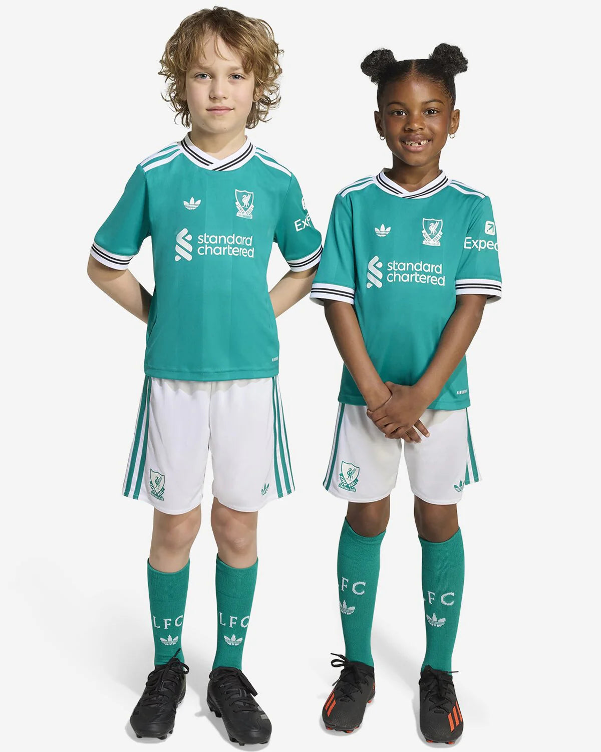 JV6470_8_APPAREL_OnModel_StandardUnisexPairShot_grey_1-lowres | UAE SPORT STORE Two children in teal Liverpool FC soccer jerseys and white shorts stand side by side, showing sponsor logos on the shirts.