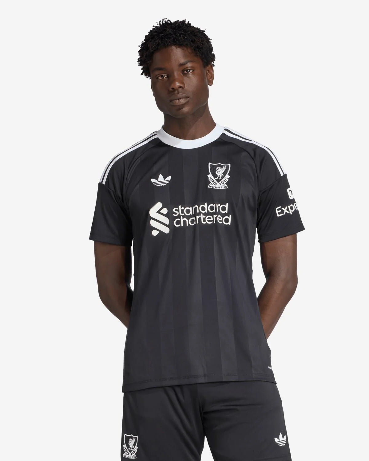 JV6477_4_APPAREL_OnModel_StandardView_custom_1-lowres A man wearing a black Liverpool FC jersey with white stripes, sponsor logo, and crest, posing against a light background.