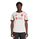 Portrait of a young Black man wearing a white Liverpool FC jersey with red crest and sponsor logos, standing against a white background, facing forward.