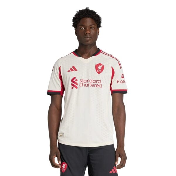Portrait of a young Black man wearing a white Liverpool FC jersey with red crest and sponsor logos, standing against a white background, facing forward.