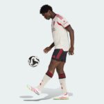 Soccer player in a white jersey and black shorts kicking a soccer ball in mid-action.