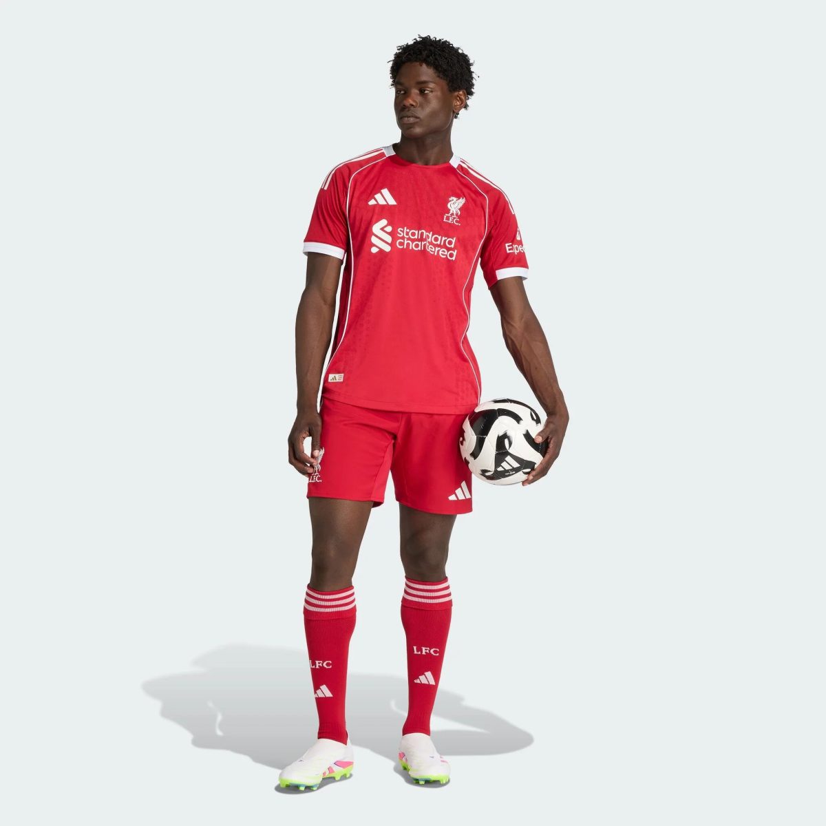 Professional male soccer player in a red Liverpool FC jersey holding a black-and-white soccer ball against a pale background, facing left.