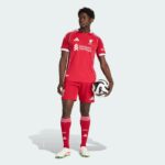 Professional male soccer player in a red Liverpool FC jersey holding a black-and-white soccer ball against a pale background, facing left.