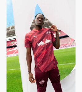 JhlBcNknPyMHZUE | UAE SPORT STORE Young man in burgundy Red Bull football kit posing in a stadium with a large white curved backdrop behind him.