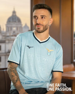 Portrait of a man in a light blue Lazio football jersey indoors, with a city skyline visible outside; slogan: ONE FAITH, ONE PASSION.