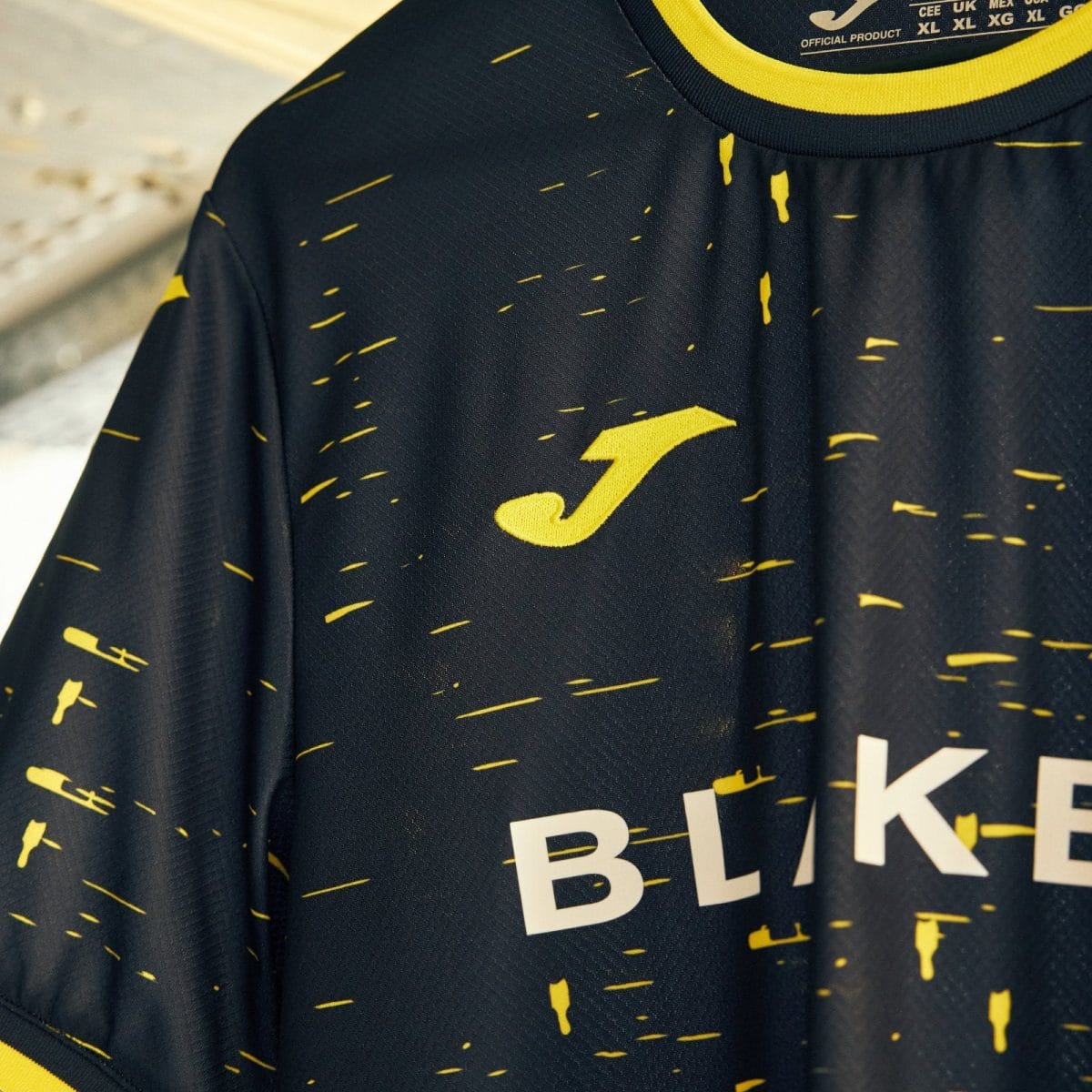 Close-up of a dark blue athletic jersey with a yellow speckle pattern and a yellow logo, with white letters 'BLK' partially visible.