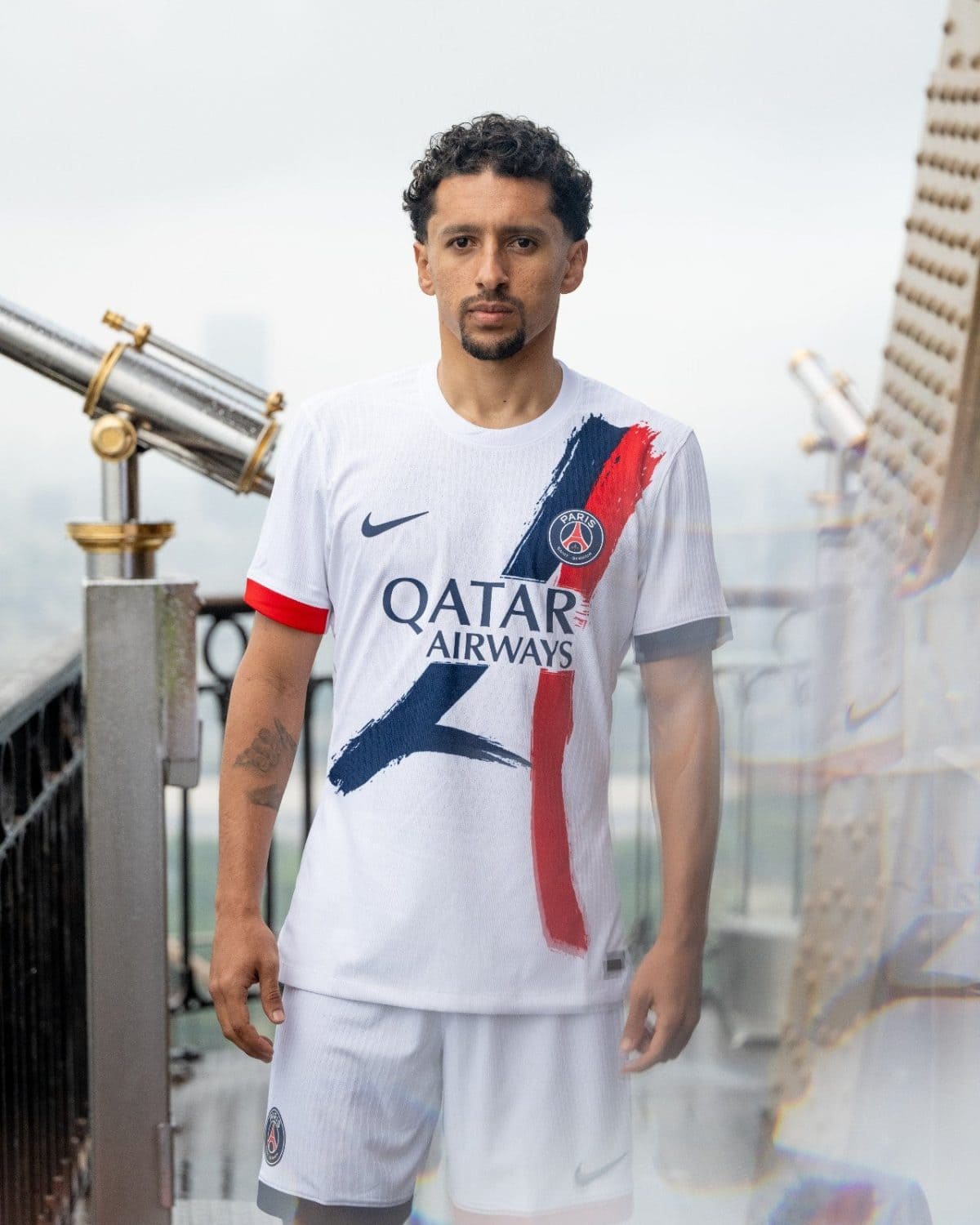 Man wearing a white Paris Saint-Germain jersey with Qatar Airways logo, standing on a rooftop with a railing and cityscape in the background.