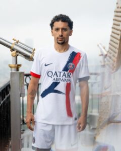 Man wearing a white Paris Saint-Germain jersey with Qatar Airways logo, standing on a rooftop with a railing and cityscape in the background.