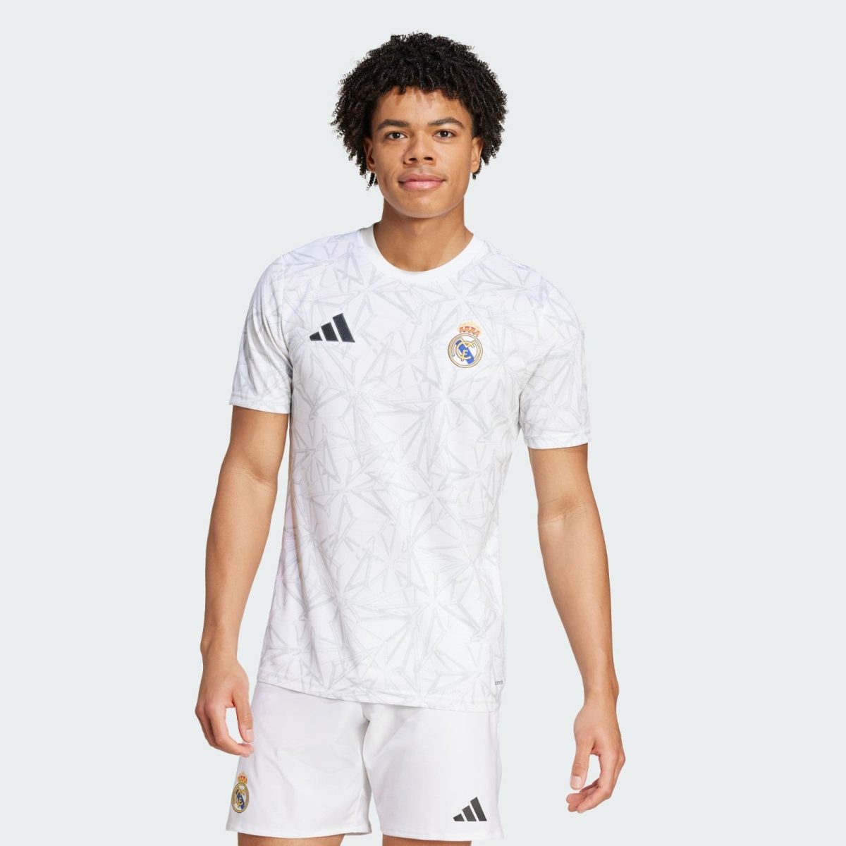 Young man modeling a white Real Madrid football jersey with Adidas logo against a pale background.