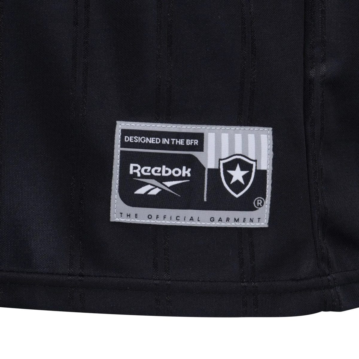 Close-up of a black garment label reading 'DESIGNED IN THE BFR' with Reebok logo and a shield/star emblem on gray stitching patch, on dark fabric.