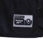 KI | UAE SPORT STORE Close-up of a black garment label reading 'DESIGNED IN THE BFR' with Reebok logo and a shield/star emblem on gray stitching patch, on dark fabric.