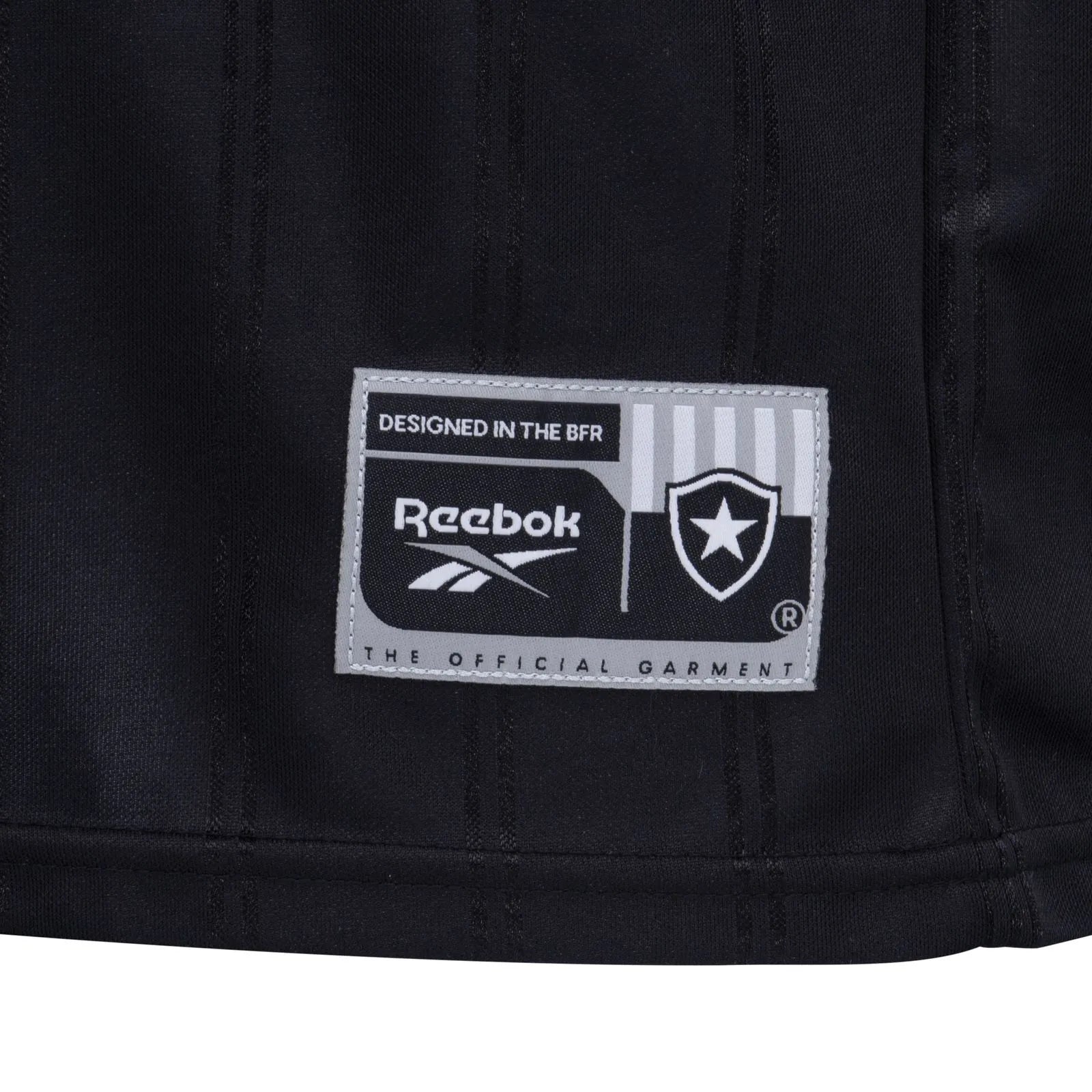 KI | UAE SPORT STORE Close-up of a black garment label reading 'DESIGNED IN THE BFR' with Reebok logo and a shield/star emblem on gray stitching patch, on dark fabric.