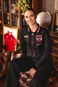 Woman in a black pinstripe jacket with an Austrian football crest, seated on a brown leather sofa in a cozy library room.