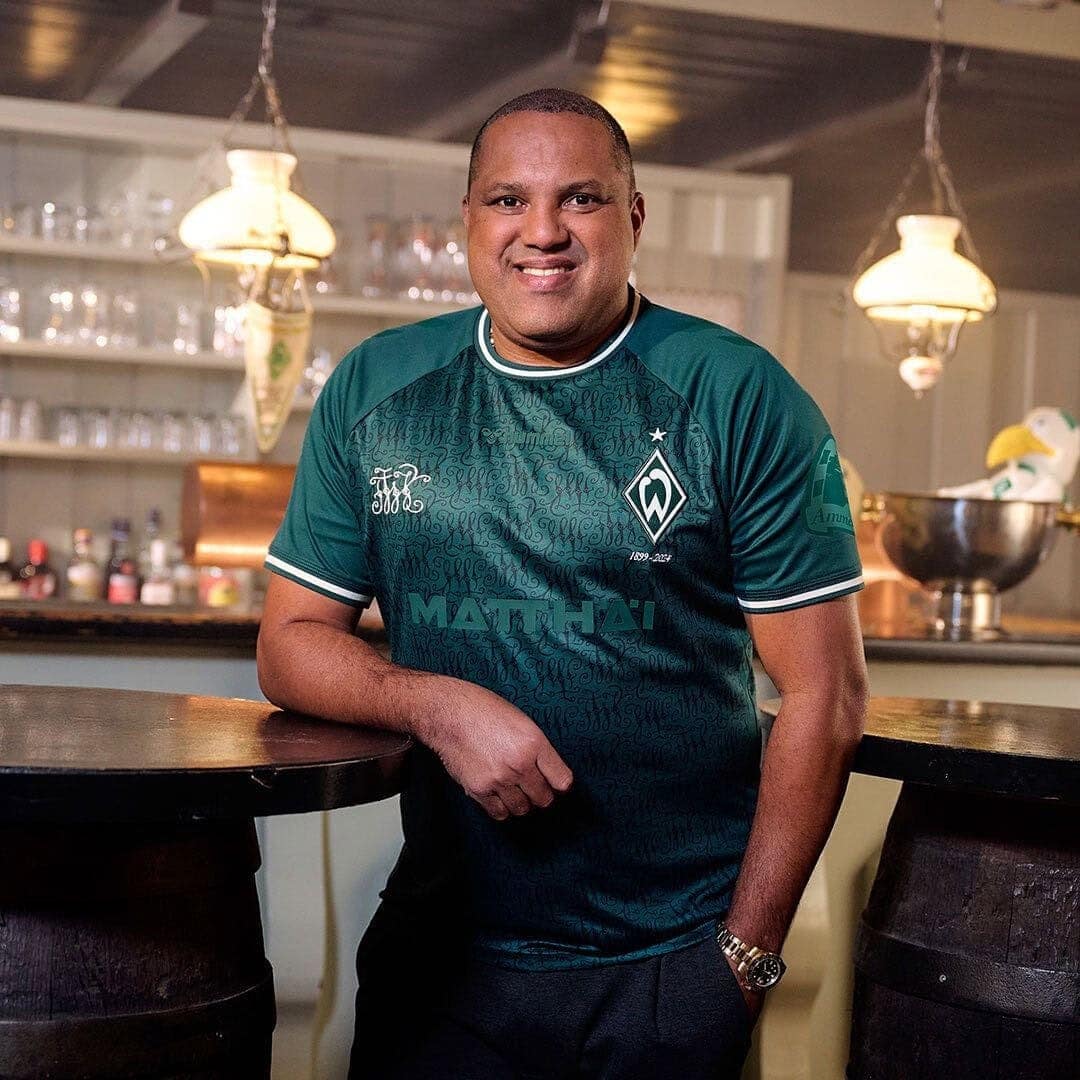 KiPhgESyOIkhiXF | UAE SPORT STORE Smiling man in a teal sports jersey leaning on a bar counter in a cozy restaurant setting.