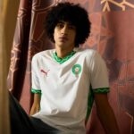 Young man with curly hair sitting against patterned curtains in a white soccer jersey with green trim and crest on chest.