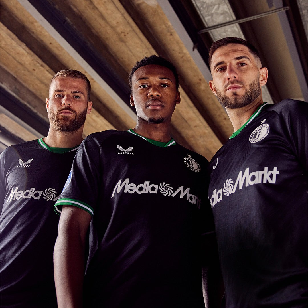 KrOsBhhrEWGRElq | UAE SPORT STORE Three male soccer players pose in black jerseys with Media Markt sponsorship under a wooden ceiling, looking at the camera.