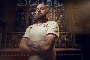 Bearded man in a white sports jersey with arms crossed, standing in a church with a stained-glass window behind him.