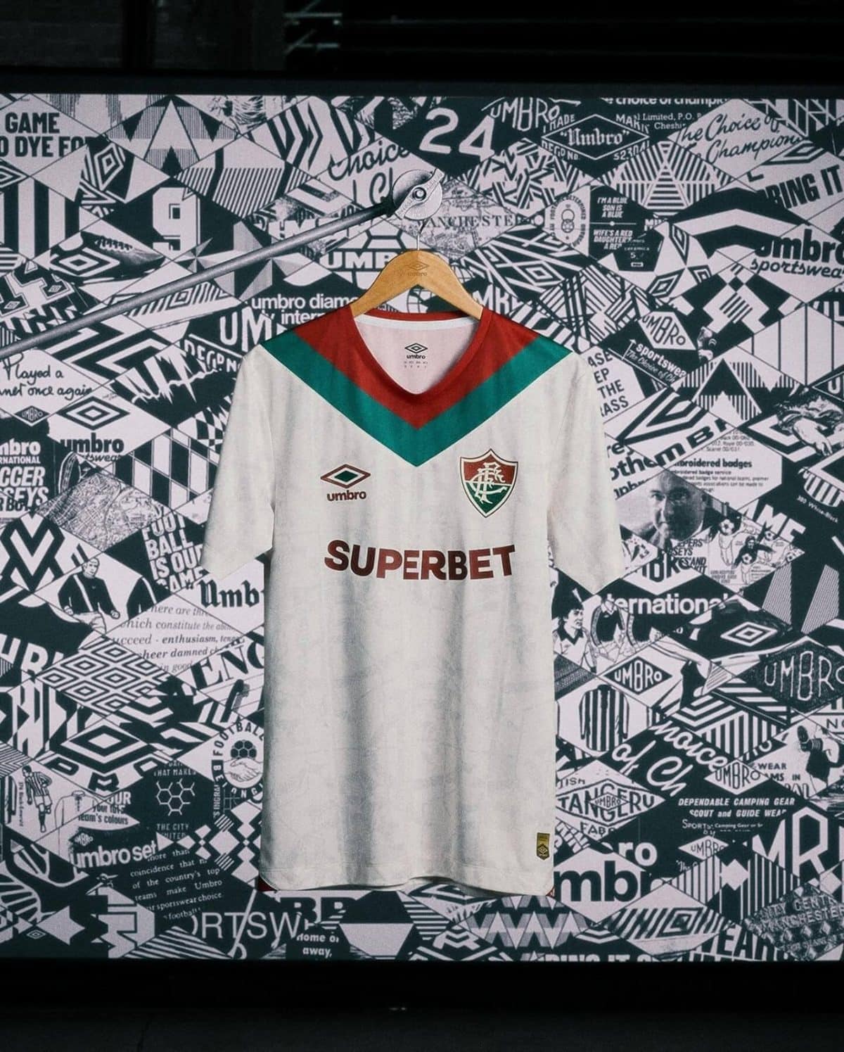 White soccer jersey on a wooden hanger, front and center, against a black-and-white graphic collage background; red V-neck with green chevron and team crest on chest.