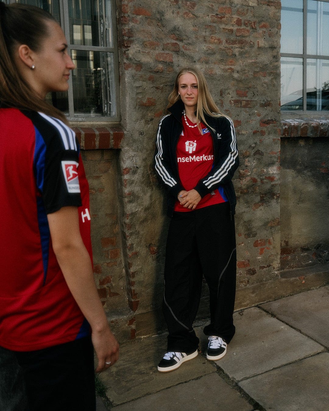 LzXHdNuBcGTij6U | UAE SPORT STORE Young woman with blonde hair in a red sports jersey and black tracksuit leaning against a brick wall, hands clasped, as another person in a red shirt stands nearby.