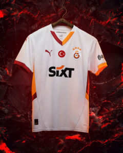 White Galatasaray football jersey with orange accents hanging on a wooden hanger against a dark red lava-like background; features SIXT sponsor, Turkish flag badge, and club crest.