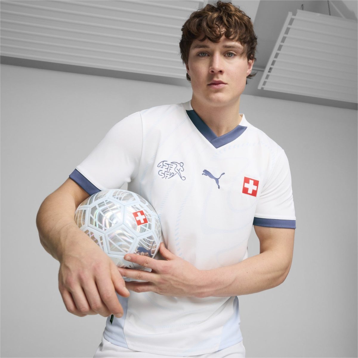 Male soccer player in white jersey holding a patterned soccer ball with a Swiss flag logo, posing in a studio.