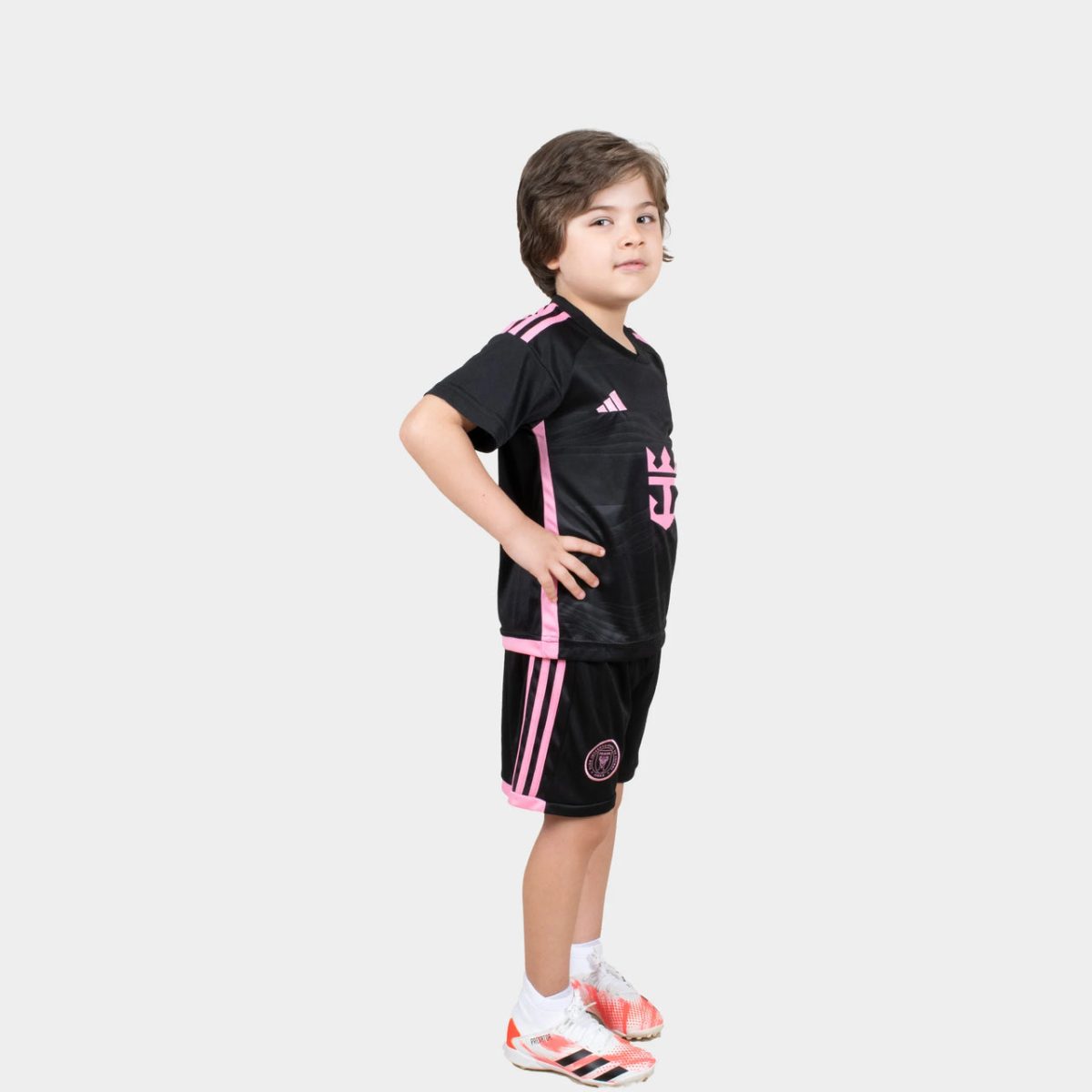 Young boy in a black and pink soccer outfit, standing in a confident side pose with hands on hips and white sneakers.