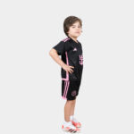 Young boy in a black and pink soccer outfit, standing in a confident side pose with hands on hips and white sneakers.