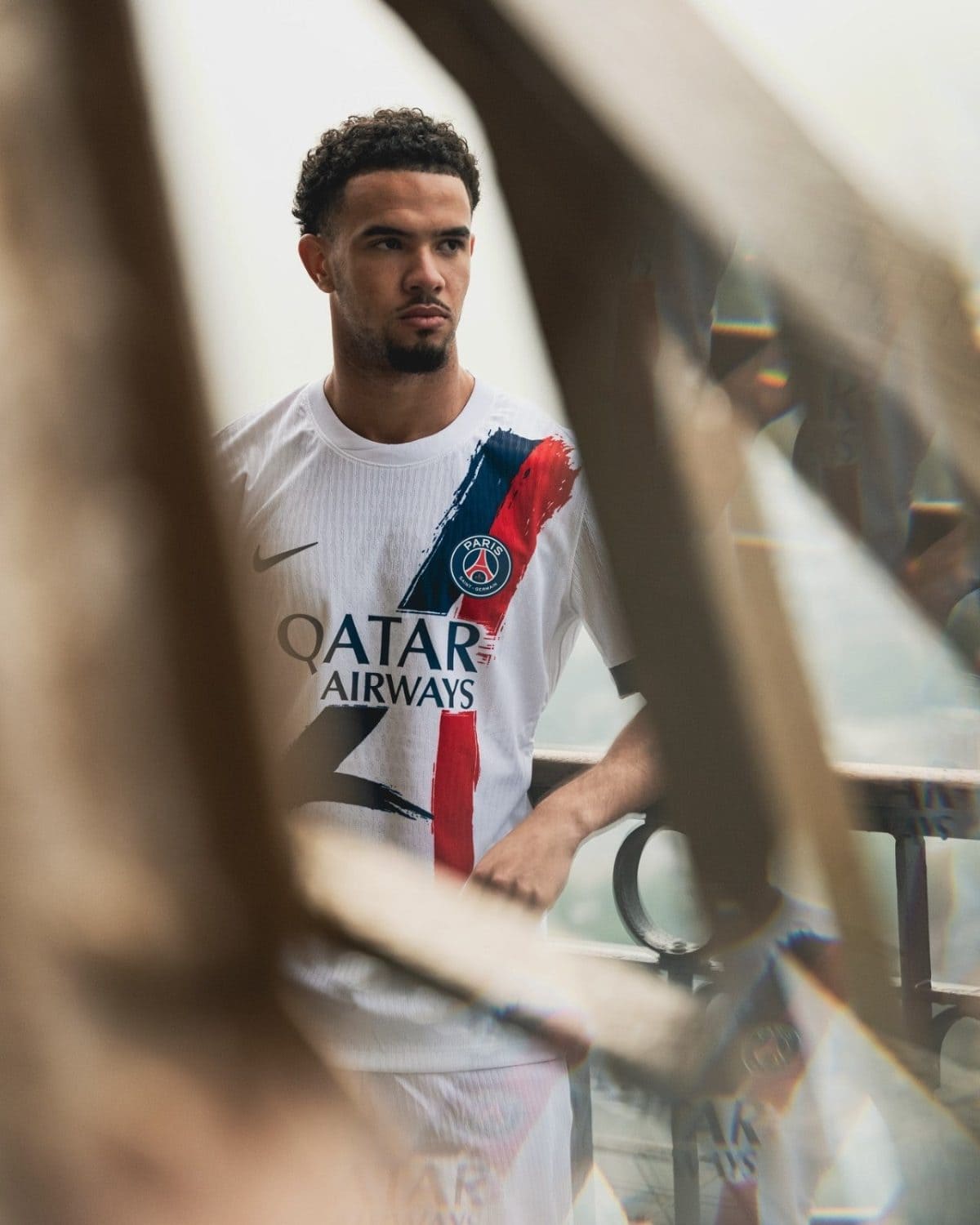 Young man in a Paris Saint-Germain jersey leaning on a railing outdoors, looking to the left.